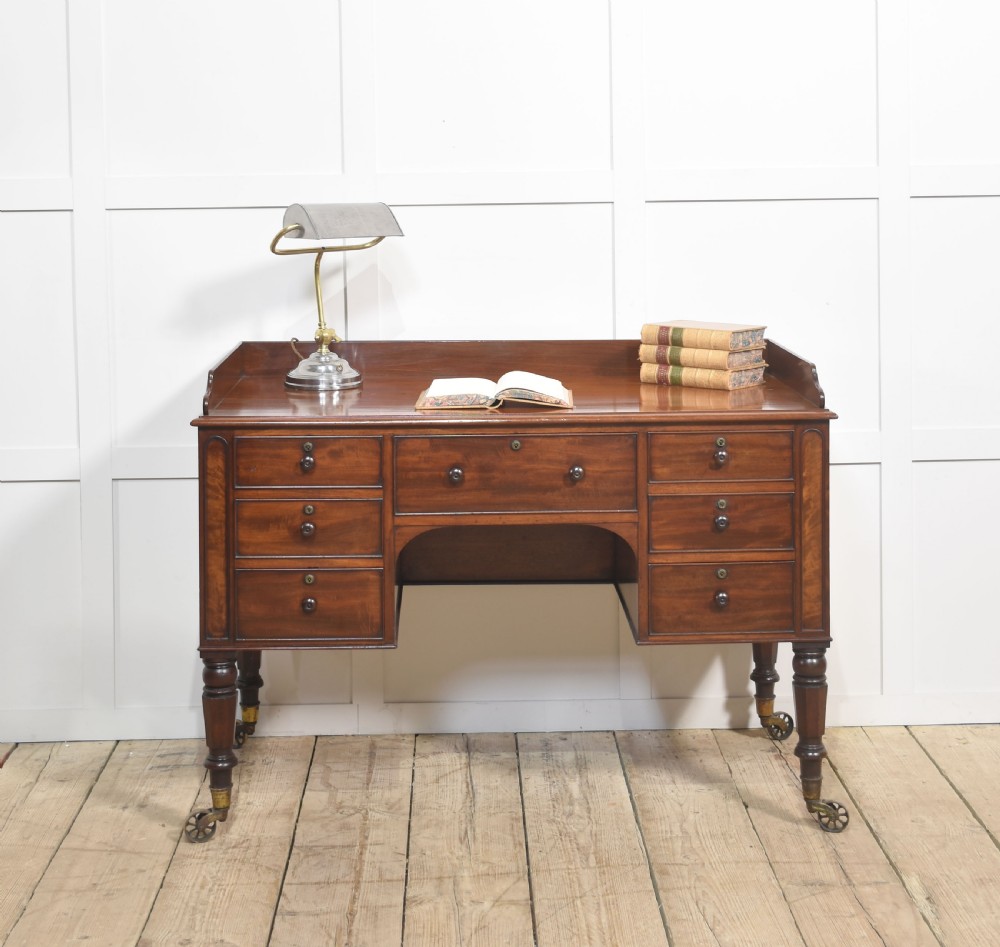 A 19th-century mahogany writing desk featuring seven drawers with dark wooden knobs, a low gallery back, turned legs, and brass castors. View V