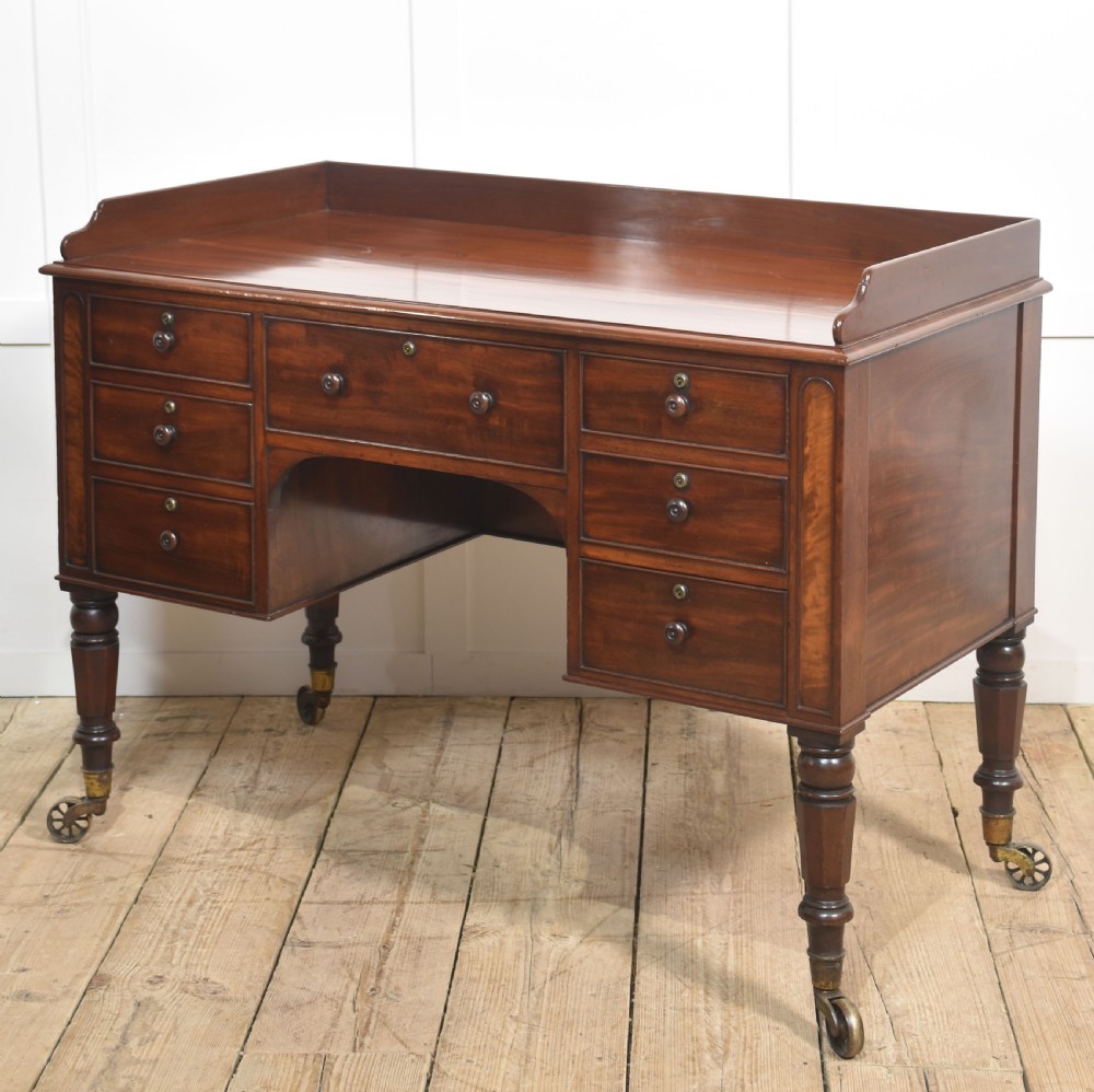 A 19th-century mahogany writing desk featuring seven drawers with dark wooden knobs, a low gallery back, turned legs, and brass castors. View VI