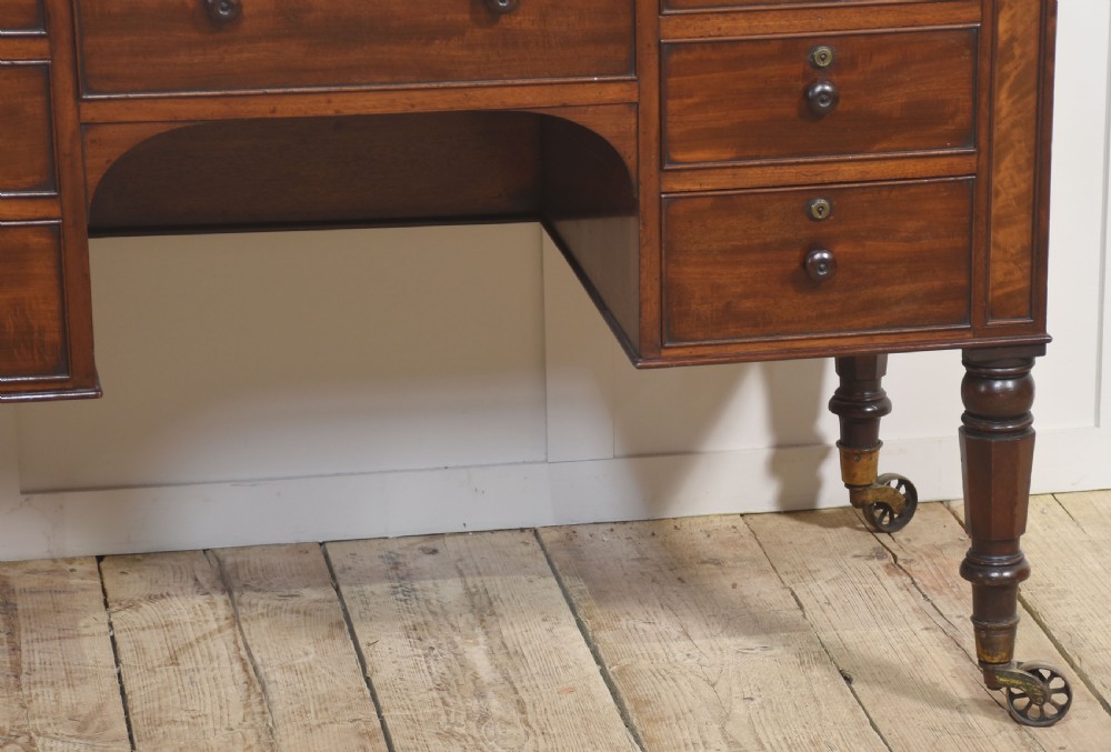 A 19th-century mahogany writing desk featuring seven drawers with dark wooden knobs, a low gallery back, turned legs, and brass castors. View VII