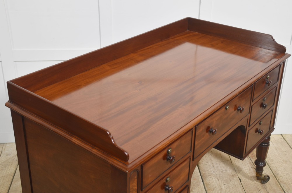 A 19th-century mahogany writing desk featuring seven drawers with dark wooden knobs, a low gallery back, turned legs, and brass castors. View XII