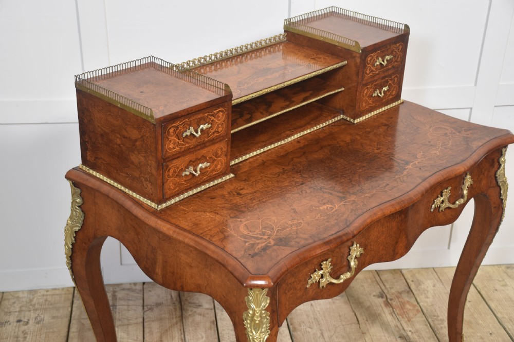A 19th-century French writing desk crafted from figured walnut wood featuring gilt-bronze ormolu mounts, cabriole legs, floral marquetry drawer fronts, and an upper superstructure with pigeonholes. View VI