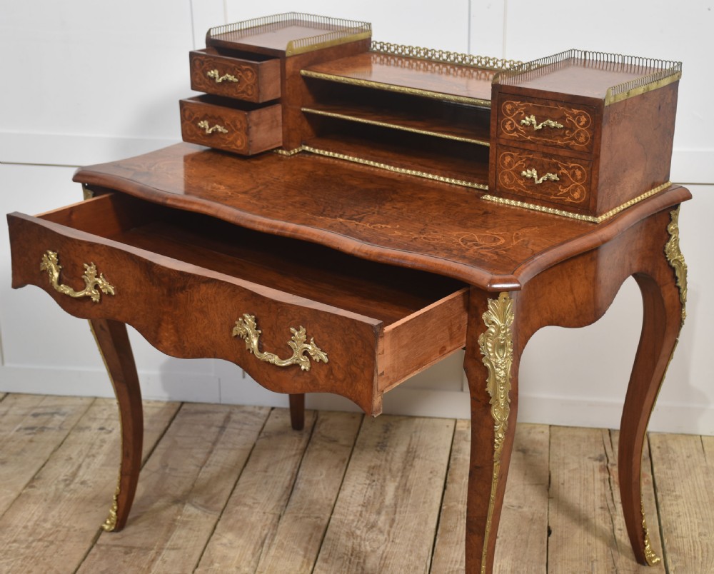 A 19th-century French writing desk crafted from figured walnut wood featuring gilt-bronze ormolu mounts, cabriole legs, floral marquetry drawer fronts, and an upper superstructure with pigeonholes. View XV