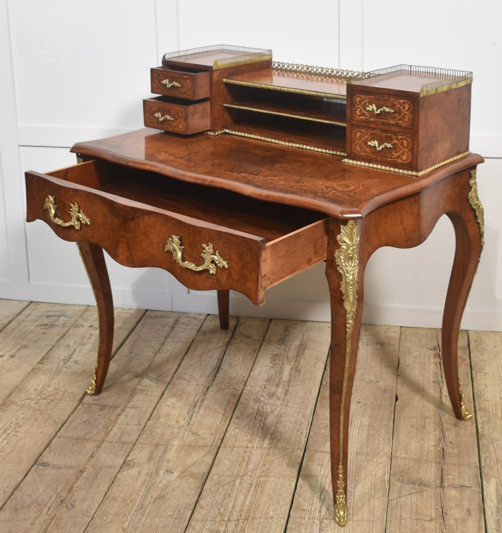 A 19th-century French writing desk crafted from figured walnut wood featuring gilt-bronze ormolu mounts, cabriole legs, floral marquetry drawer fronts, and an upper superstructure with pigeonholes. View XVI
