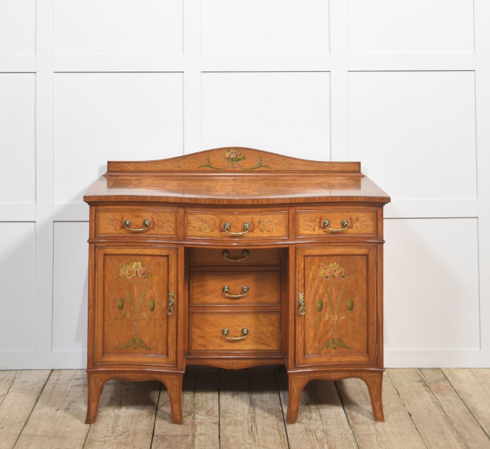 An antique painted satinwood dressing table featuring floral artwork, a central kneehole, side cupboards, drawers, brass handles, and a low gallery back.