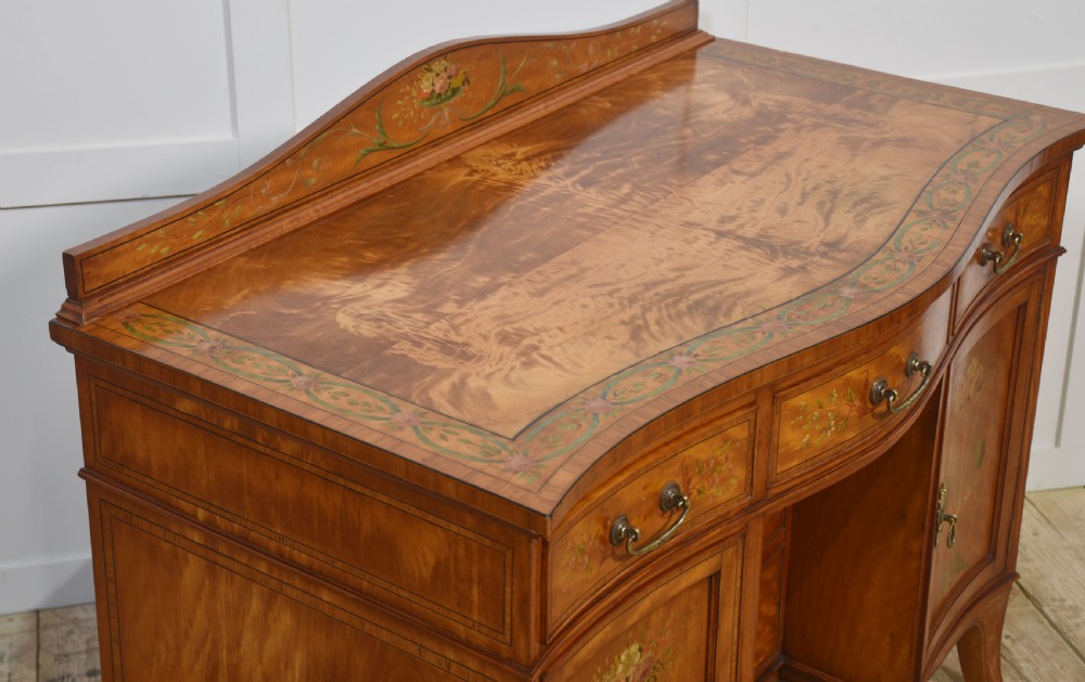 An antique painted satinwood dressing table featuring floral artwork, a central kneehole, side cupboards, drawers, brass handles, and a low gallery back. View X