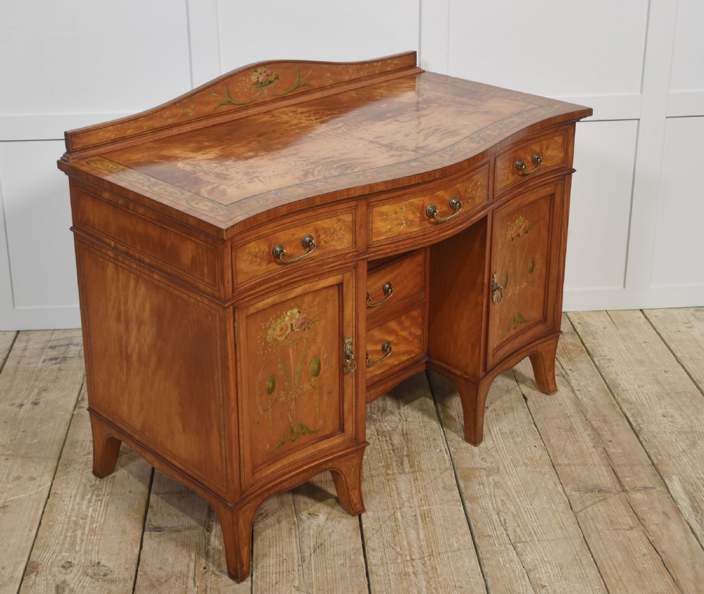 An antique painted satinwood dressing table featuring floral artwork, a central kneehole, side cupboards, drawers, brass handles, and a low gallery back. View XII