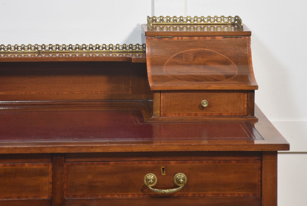 An antique mahogany writing desk featuring satinwood inlay, brass gallery rails, two frieze drawers, and square tapered legs on castors. View II