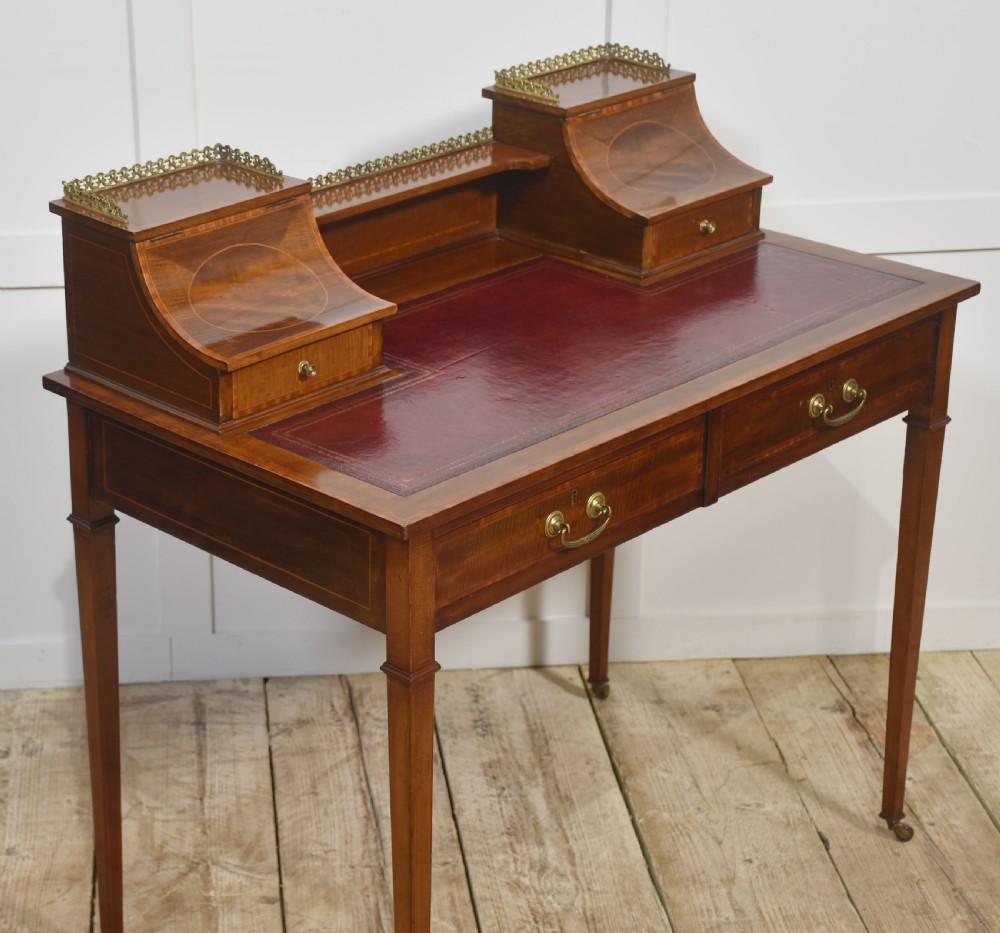 An antique mahogany writing desk featuring satinwood inlay, brass gallery rails, two frieze drawers, and square tapered legs on castors. View III