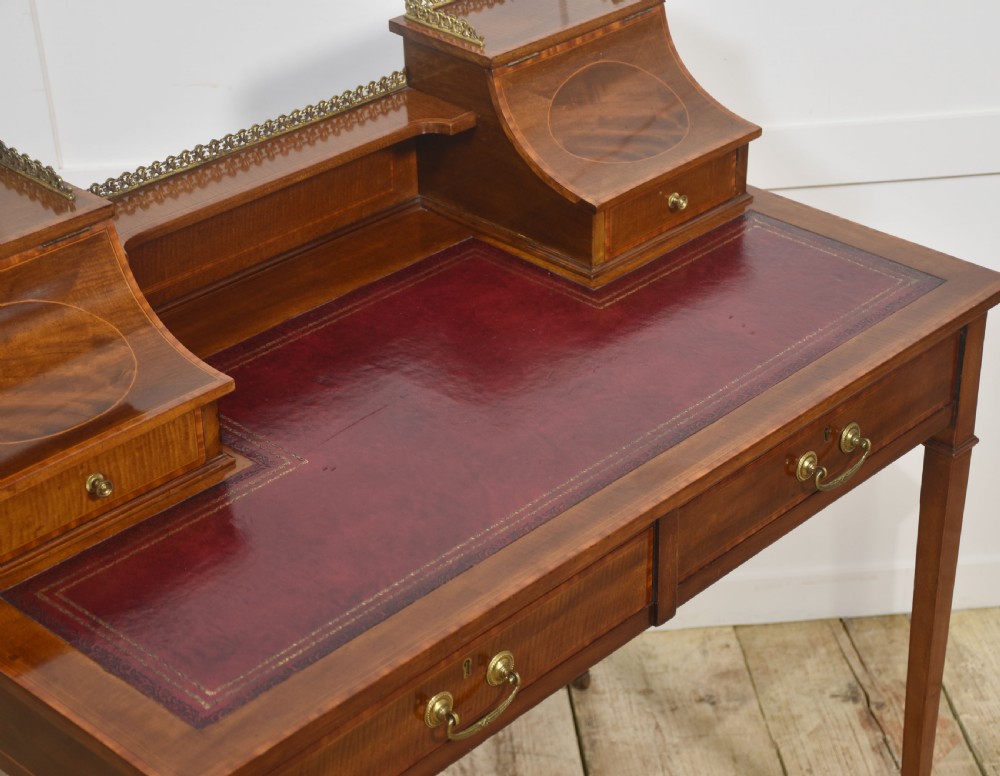 An antique mahogany writing desk featuring satinwood inlay, brass gallery rails, two frieze drawers, and square tapered legs on castors. View VII