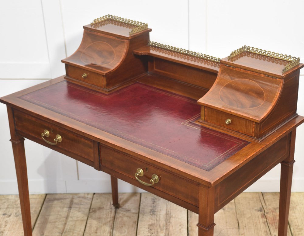 An antique mahogany writing desk featuring satinwood inlay, brass gallery rails, two frieze drawers, and square tapered legs on castors. View VIII