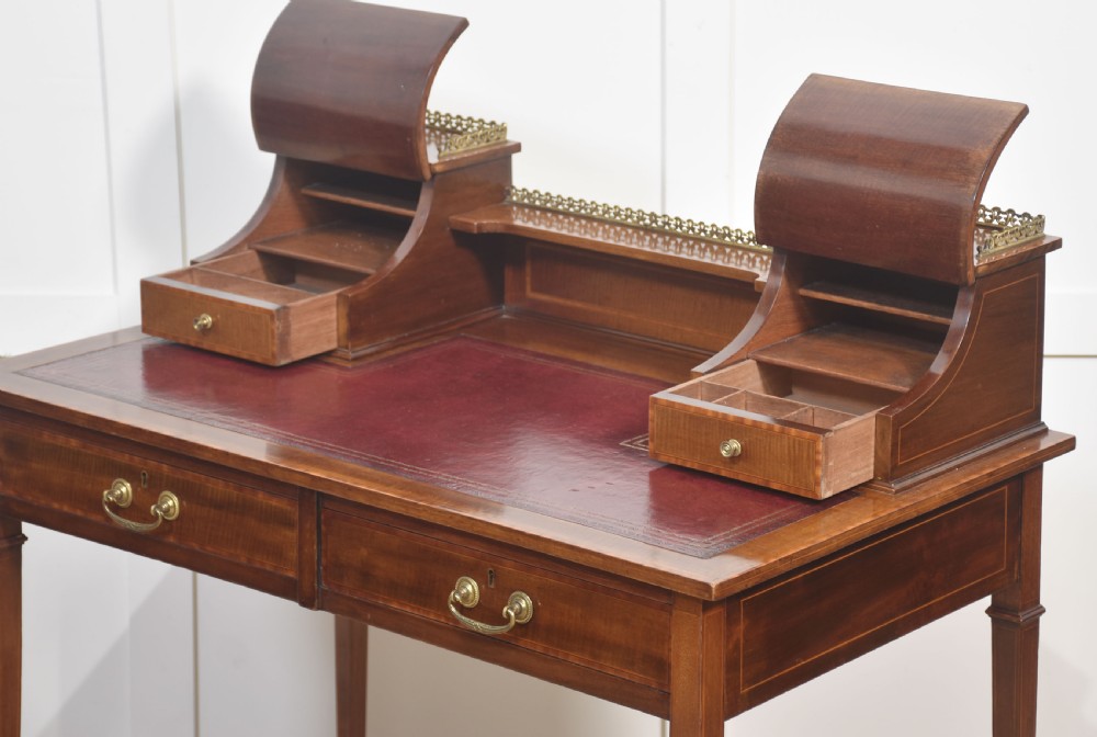 An antique mahogany writing desk featuring satinwood inlay, brass gallery rails, two frieze drawers, and square tapered legs on castors. View X
