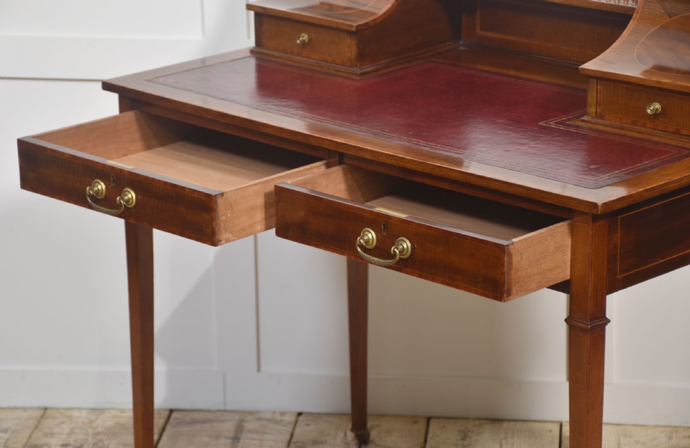 An antique mahogany writing desk featuring satinwood inlay, brass gallery rails, two frieze drawers, and square tapered legs on castors. View XII