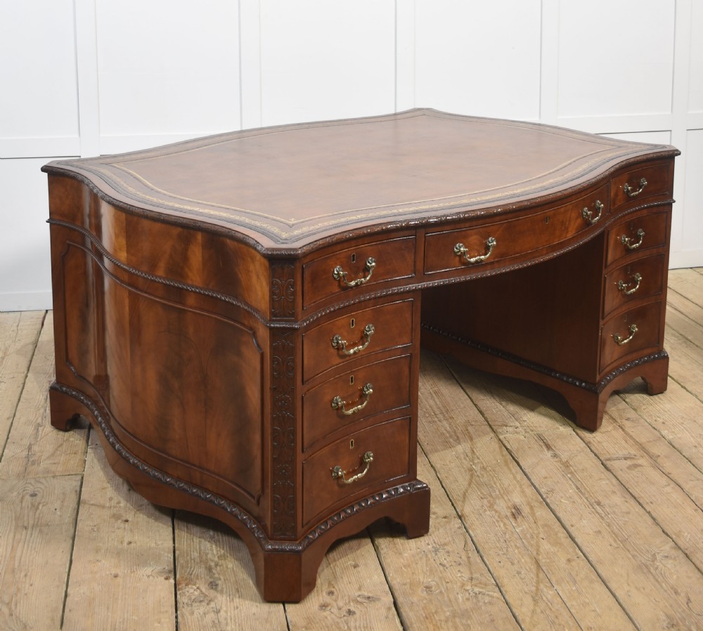 A large Chippendale Revival mahogany partners desk with a serpentine front, gilded leather inset writing top, and brass swan-neck handles.