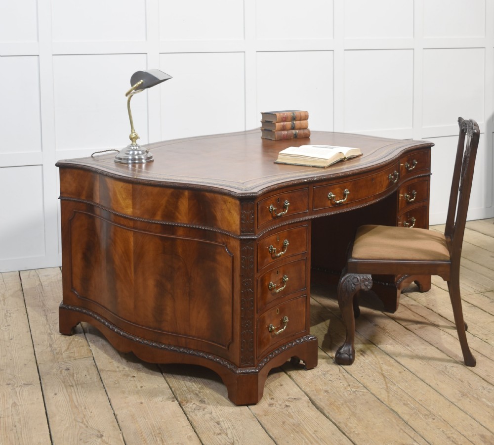 A large Chippendale Revival mahogany partners desk with a serpentine front, gilded leather inset writing top, and brass swan-neck handles. View VI