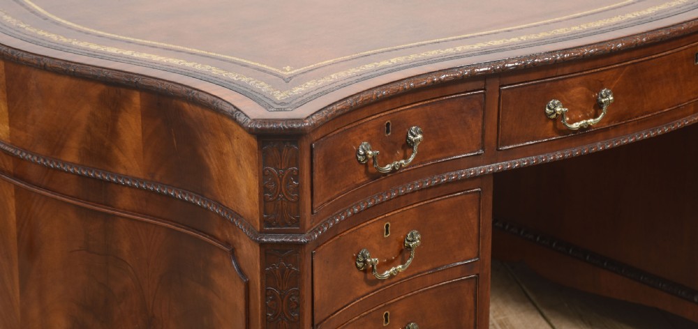 A large Chippendale Revival mahogany partners desk with a serpentine front, gilded leather inset writing top, and brass swan-neck handles. View IX