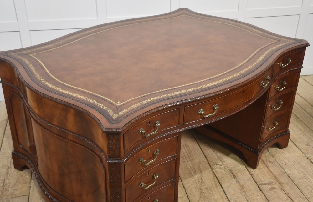 A large Chippendale Revival mahogany partners desk with a serpentine front, gilded leather inset writing top, and brass swan-neck handles. View X