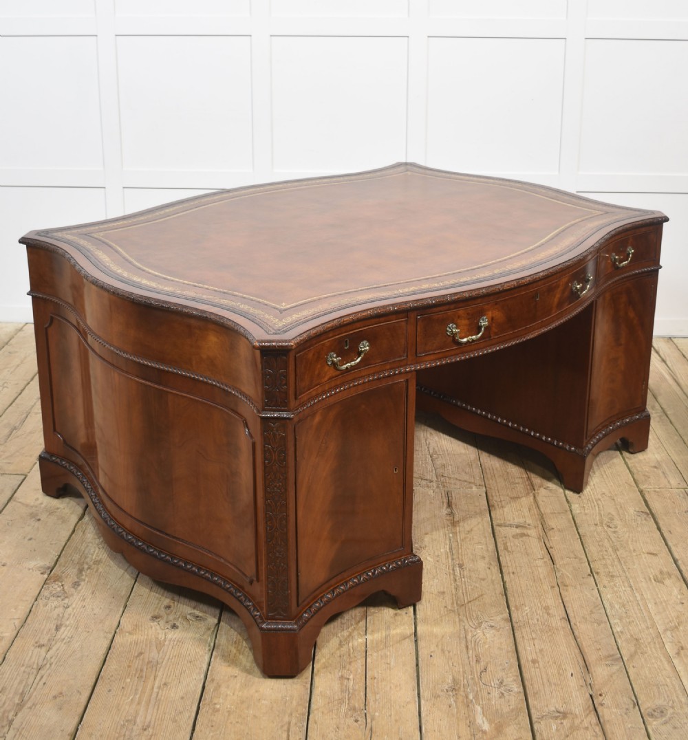 A large Chippendale Revival mahogany partners desk with a serpentine front, gilded leather inset writing top, and brass swan-neck handles. View XVII