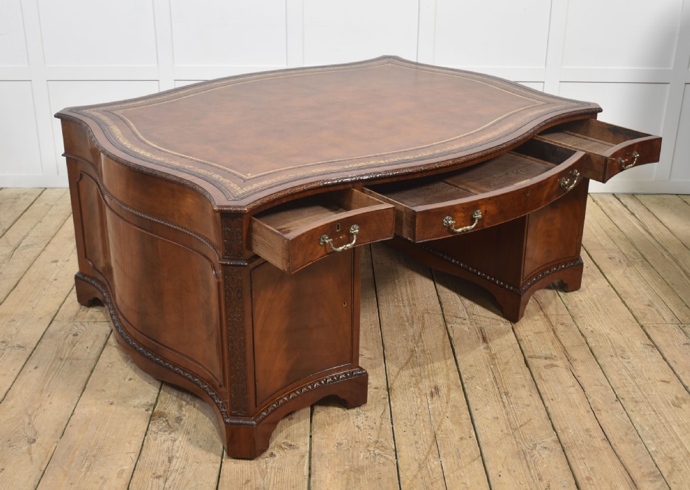 A large Chippendale Revival mahogany partners desk with a serpentine front, gilded leather inset writing top, and brass swan-neck handles. View XIX