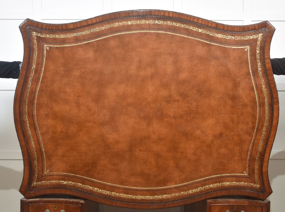 A large Chippendale Revival mahogany partners desk with a serpentine front, gilded leather inset writing top, and brass swan-neck handles. View XX
