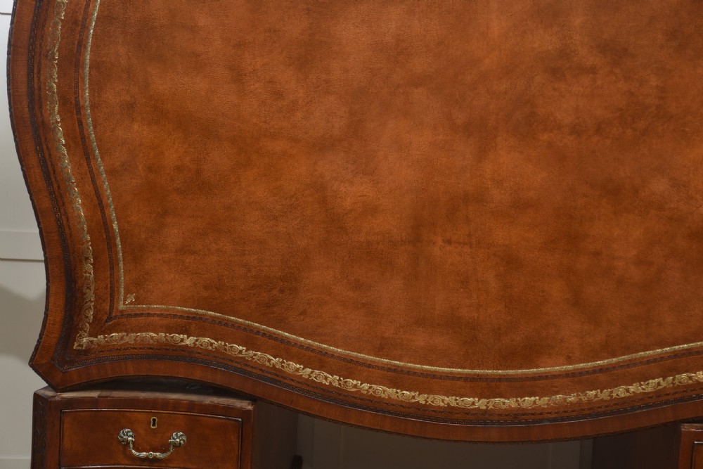 A large Chippendale Revival mahogany partners desk with a serpentine front, gilded leather inset writing top, and brass swan-neck handles. View XXI