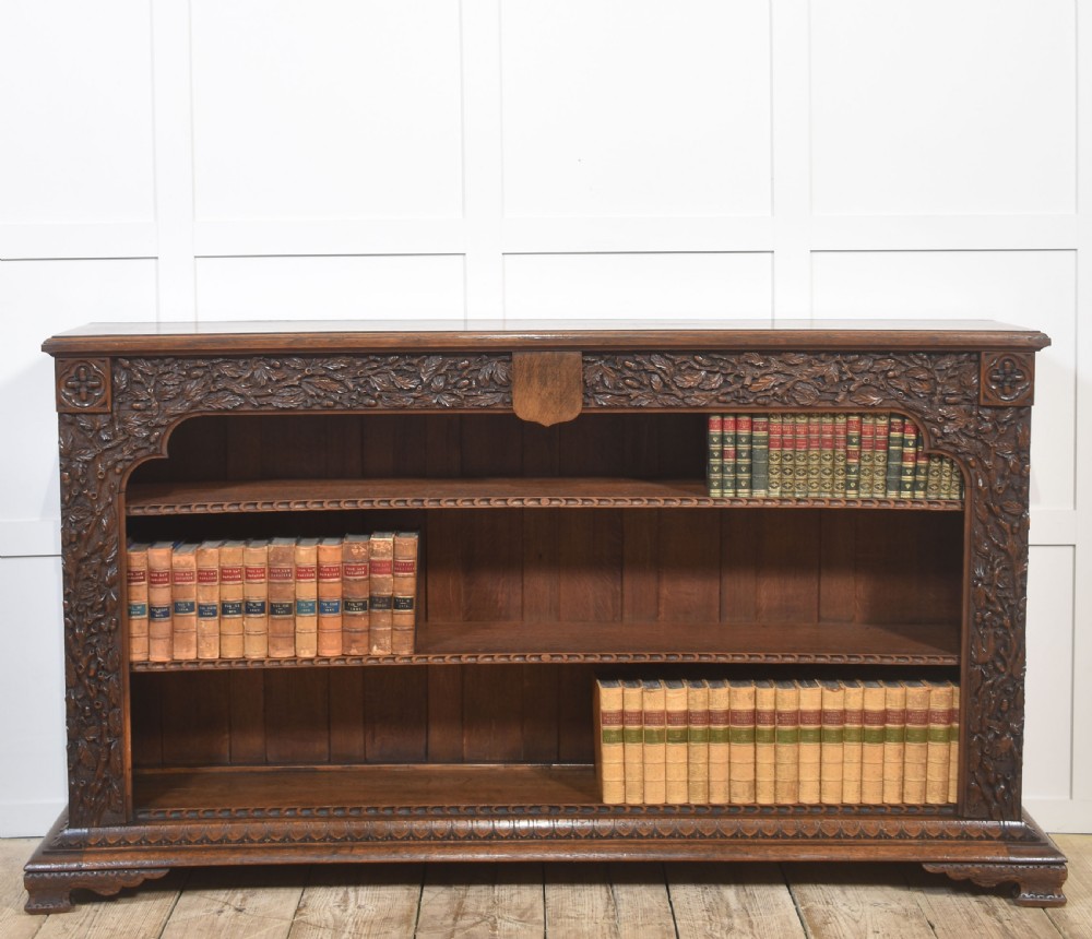 A low carved dark oak open bookcase featuring three wide shelves flanked by carved foliate borders and a central shield motif.