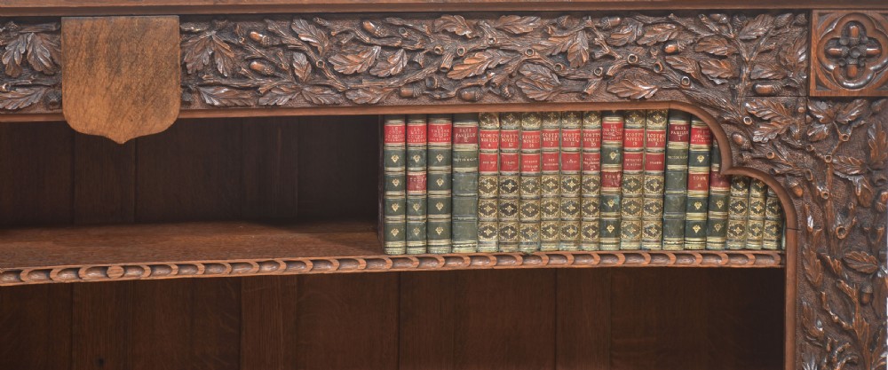 A low carved dark oak open bookcase featuring three wide shelves flanked by carved foliate borders and a central shield motif. View II