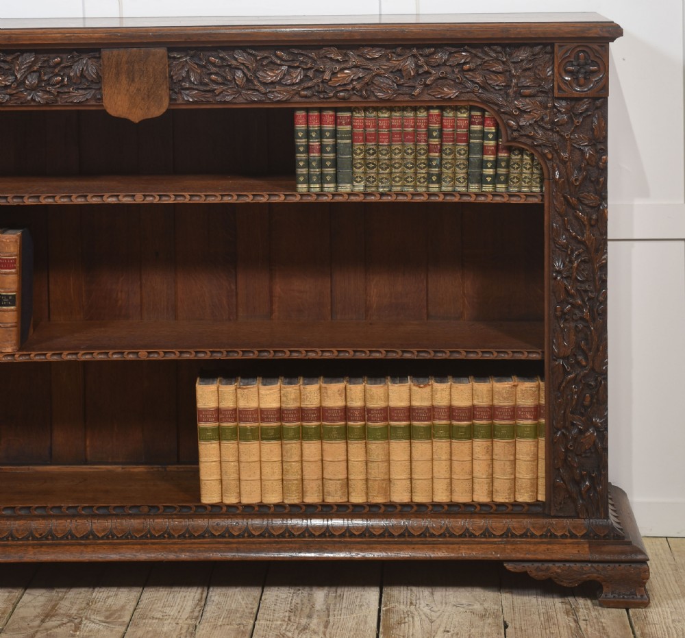 A low carved dark oak open bookcase featuring three wide shelves flanked by carved foliate borders and a central shield motif. View III