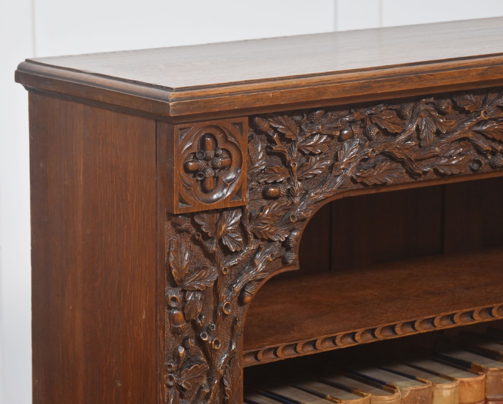 A low carved dark oak open bookcase featuring three wide shelves flanked by carved foliate borders and a central shield motif. View V