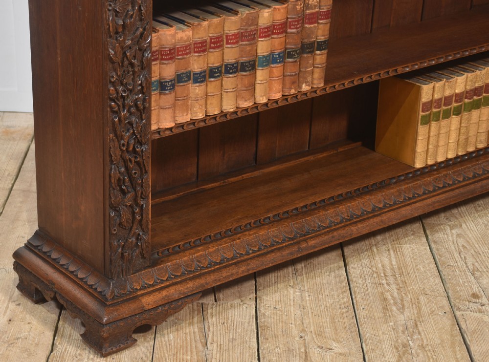 A low carved dark oak open bookcase featuring three wide shelves flanked by carved foliate borders and a central shield motif. View VI