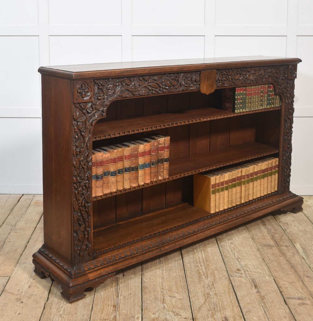 A low carved dark oak open bookcase featuring three wide shelves flanked by carved foliate borders and a central shield motif. View VII