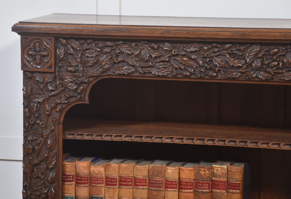 A low carved dark oak open bookcase featuring three wide shelves flanked by carved foliate borders and a central shield motif. View VIII