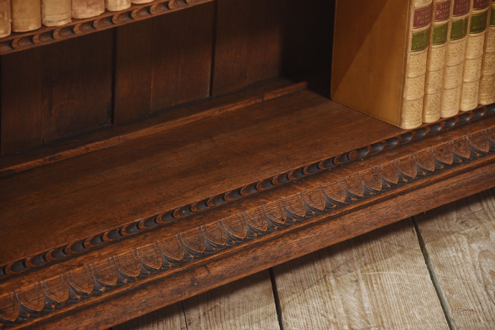 A low carved dark oak open bookcase featuring three wide shelves flanked by carved foliate borders and a central shield motif. View X