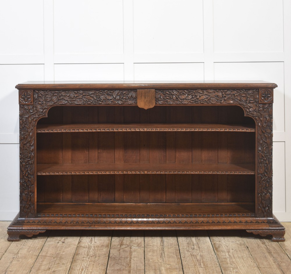 A low carved dark oak open bookcase featuring three wide shelves flanked by carved foliate borders and a central shield motif. View XI