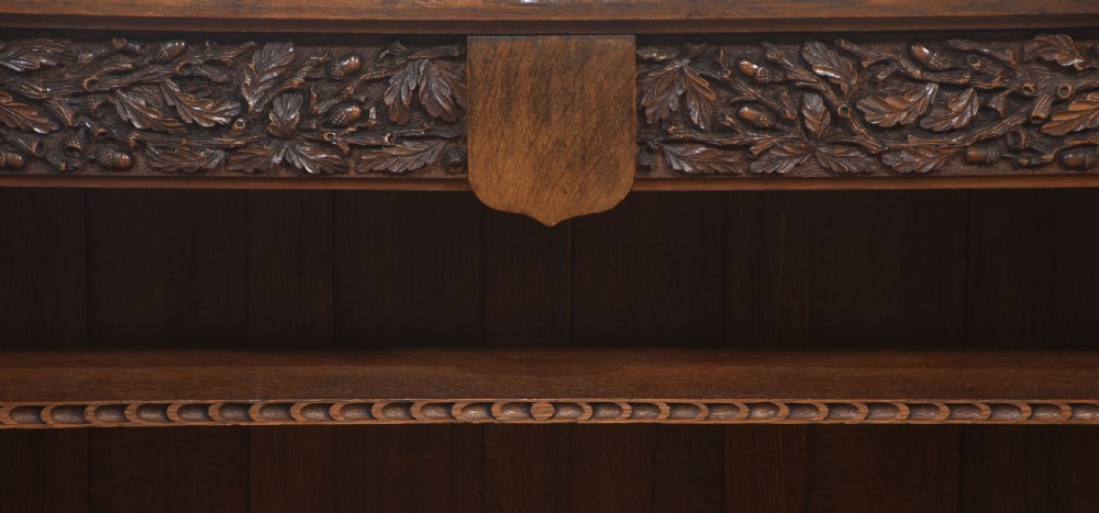 A low carved dark oak open bookcase featuring three wide shelves flanked by carved foliate borders and a central shield motif. View XII