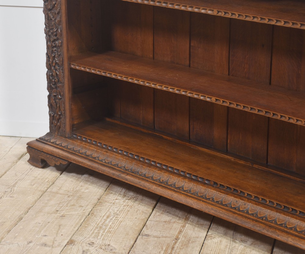 A low carved dark oak open bookcase featuring three wide shelves flanked by carved foliate borders and a central shield motif. View XV