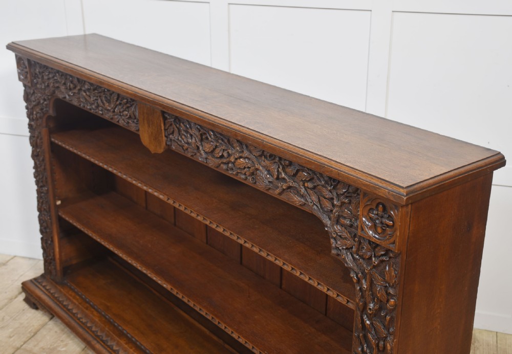 A low carved dark oak open bookcase featuring three wide shelves flanked by carved foliate borders and a central shield motif. View XVI