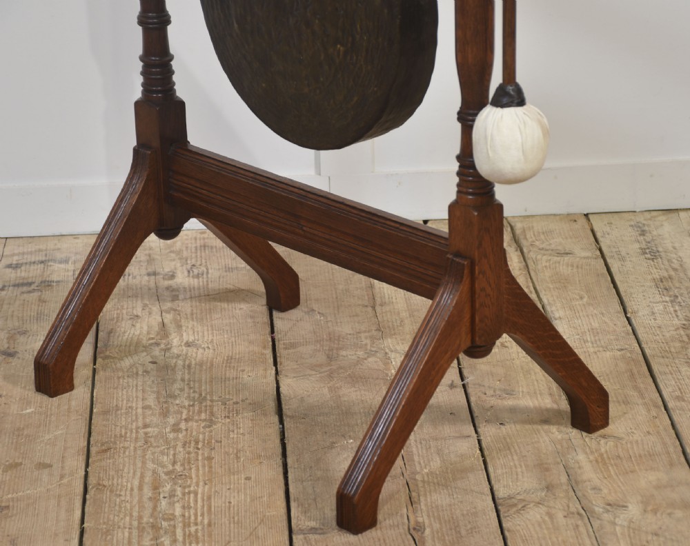 An antique dark oak floor-standing dinner gong featuring turned upright supports, top finials, a suspended circular metal disc, and an attached padded striker. View V