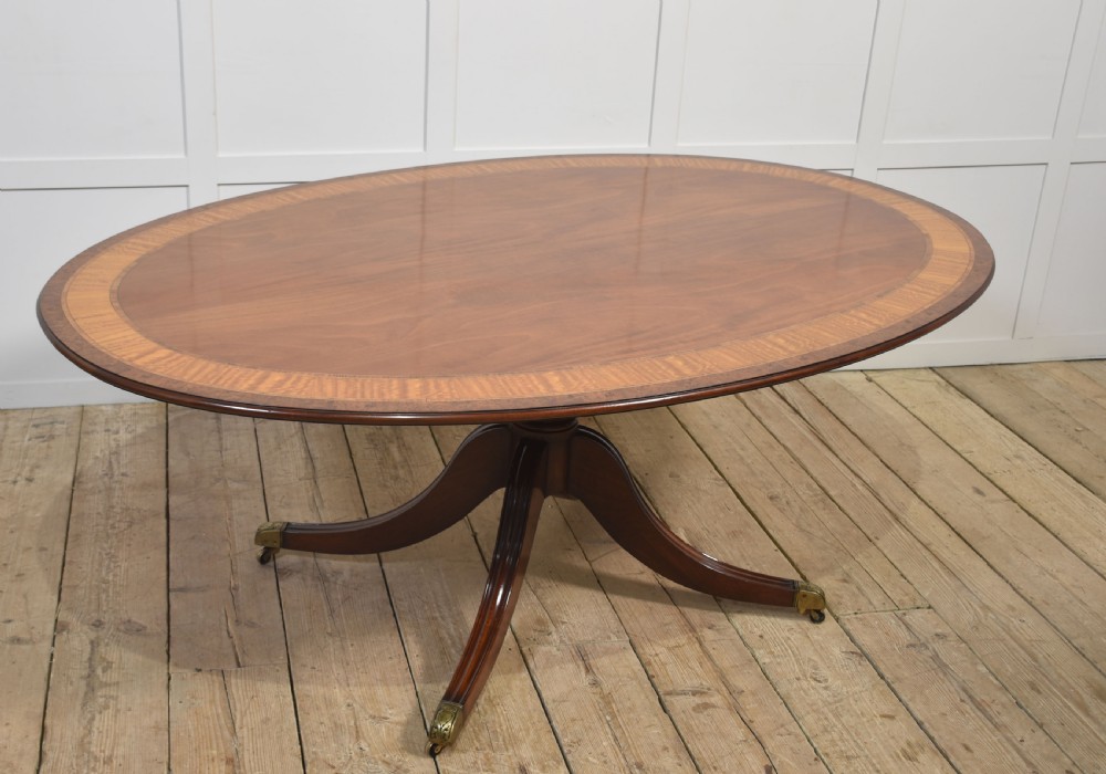 An oval mahogany dining table featuring a crossbanded satinwood border and a central pedestal with four reeded splayed legs ending in brass cap castors. View II