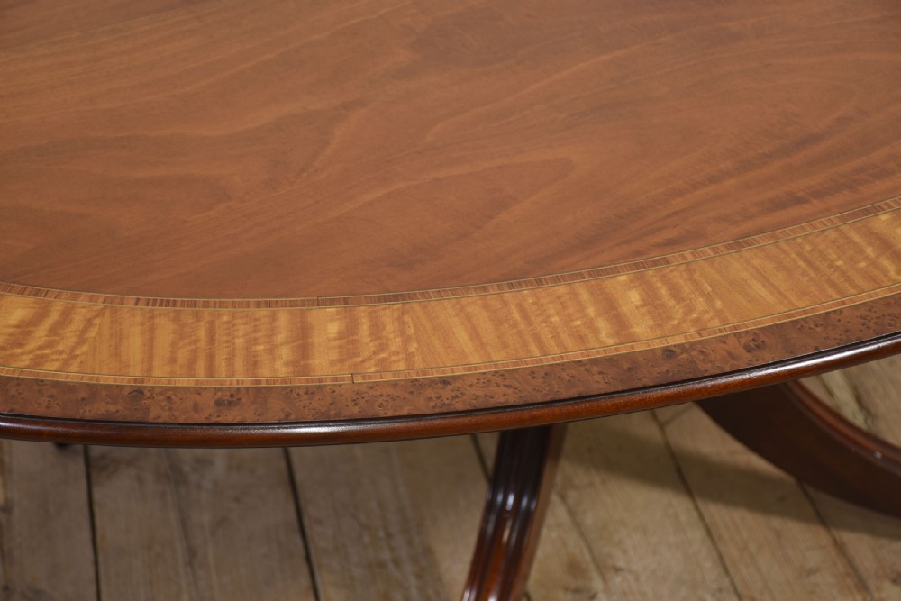An oval mahogany dining table featuring a crossbanded satinwood border and a central pedestal with four reeded splayed legs ending in brass cap castors. View IV