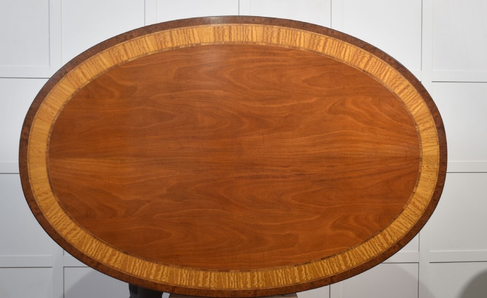 An oval mahogany dining table featuring a crossbanded satinwood border and a central pedestal with four reeded splayed legs ending in brass cap castors. View V