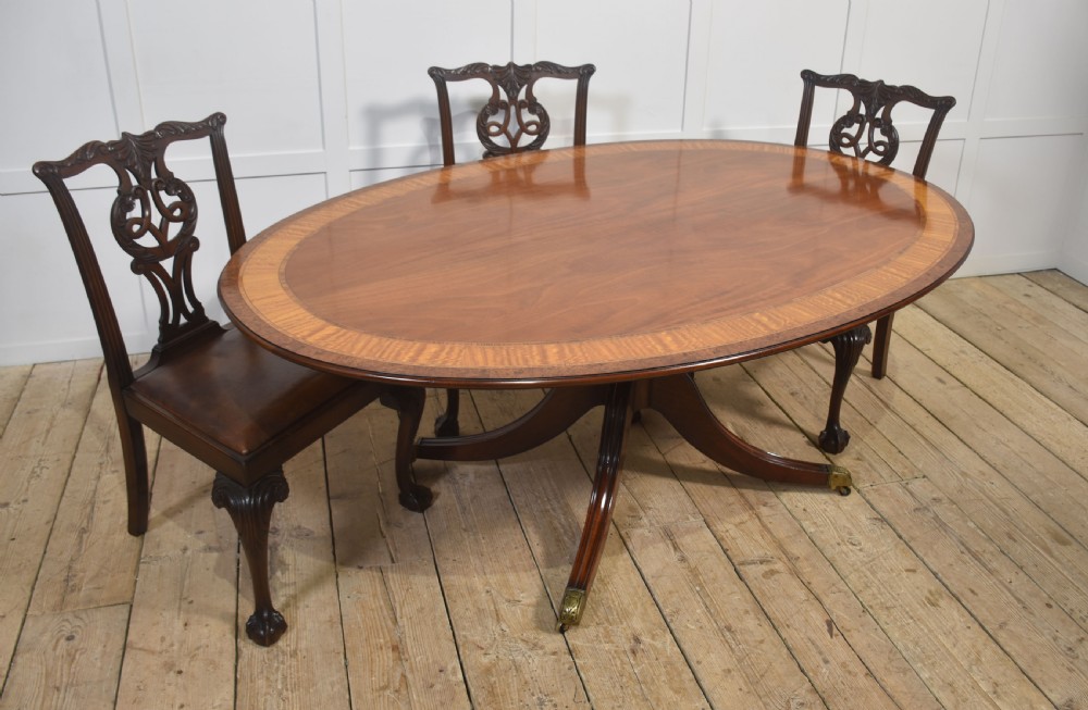 An oval mahogany dining table featuring a crossbanded satinwood border and a central pedestal with four reeded splayed legs ending in brass cap castors. View VI