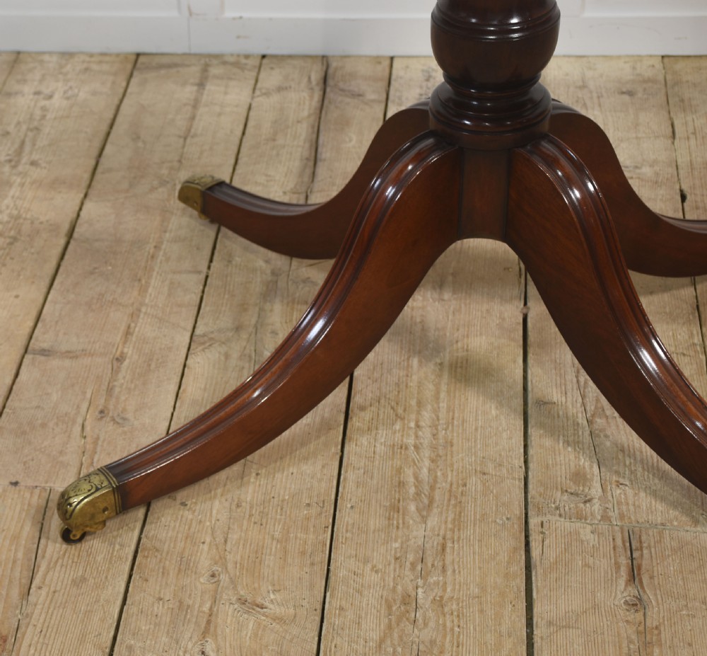 An oval mahogany dining table featuring a crossbanded satinwood border and a central pedestal with four reeded splayed legs ending in brass cap castors. View VII