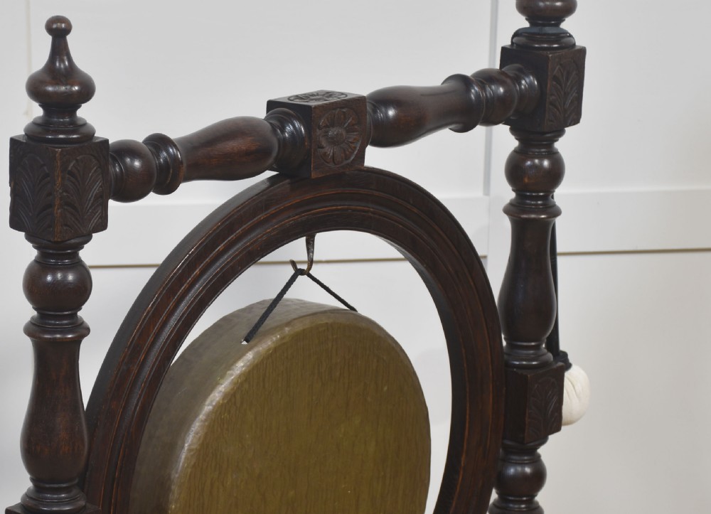 A freestanding dark oak frame with carved posts, turned finials, and shoe feet, housing a suspended circular brass gong with a matching beater attached at the side. View V