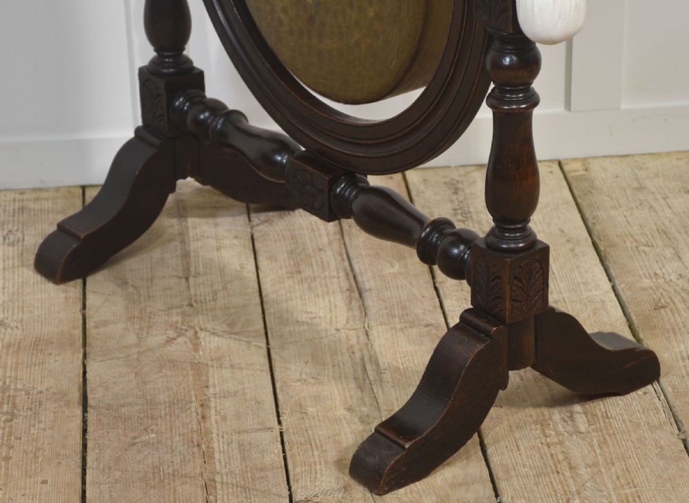 A freestanding dark oak frame with carved posts, turned finials, and shoe feet, housing a suspended circular brass gong with a matching beater attached at the side. View IX