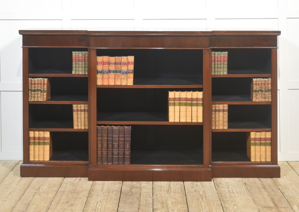 A wide mahogany open breakfront bookcase with four tiers of shelving containing antique leather-bound books.