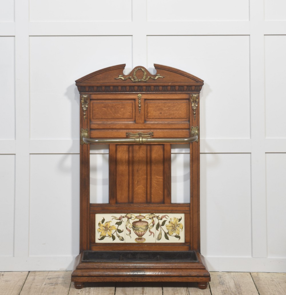 An antique oak umbrella stand featuring an arched top pediment, brass retaining rail, decorative ceramic floral tiles, and a lower drip tray.