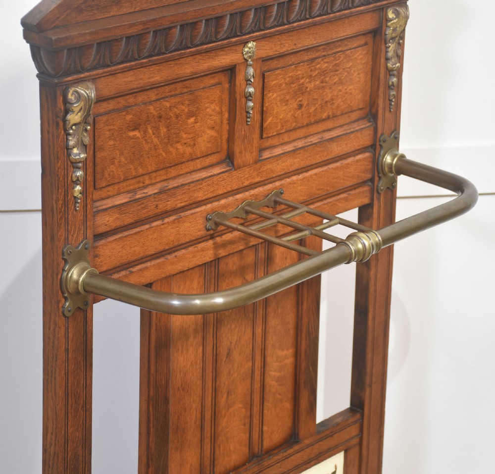An antique oak umbrella stand featuring an arched top pediment, brass retaining rail, decorative ceramic floral tiles, and a lower drip tray. View X