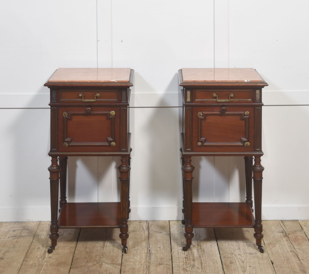 A matching pair of antique mahogany bedside cabinets with pink marble tops, single drawers, framed cupboard doors, lower shelves, and fluted legs on castors.