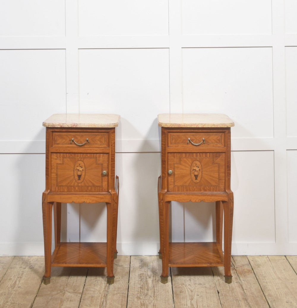 A pair of tall French satinwood bedside cabinets with marble tops, marquetry oval medallions, drawers, and slender legs.