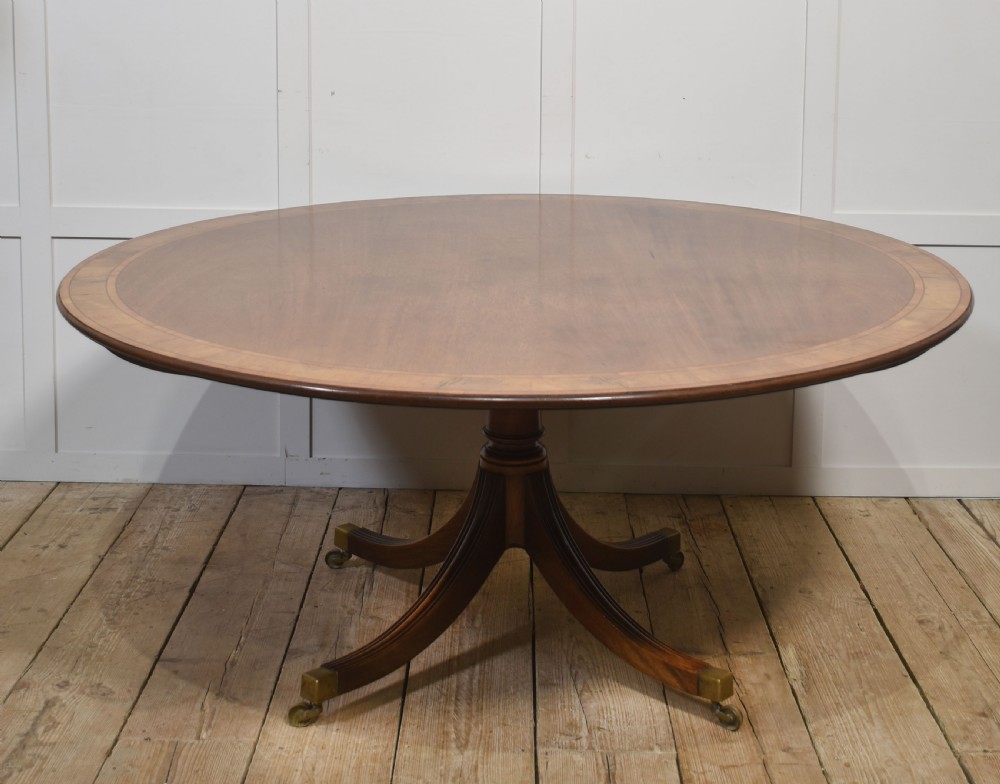 A circular Georgian-style mahogany dining table featuring a crossbanded inlay border, supported by a turned central pedestal with four reeded splayed legs on brass castors.